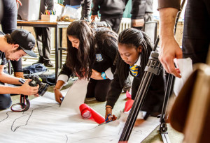 Girls cutting out a large paper figure, while being filmed