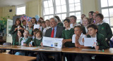 Man seated at a table with large group of schoochildren, displaying artwork.