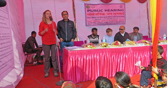 Girl speaking with microphone, standing next to podium with several people seated behind it.